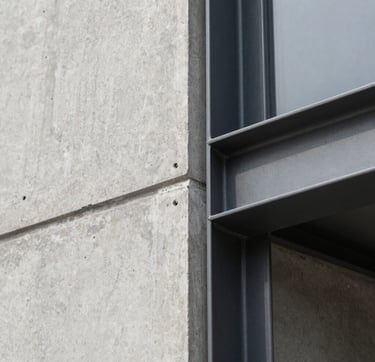 A close-up architectural detail focusing on tectonic precision. The image shows a clean joint between a light neutral gray concrete wall and a dark charcoal gray steel frame. The lighting is diffused and natural, highlighting the texture of the materials and the studio's commitment to high technical rigor.