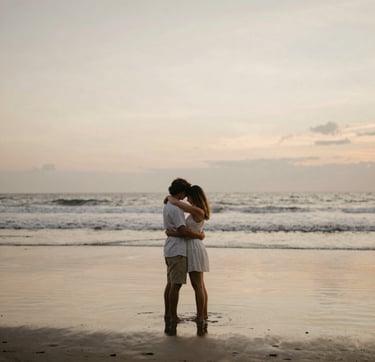 A warm, intimate wide shot of a couple embracing on a quiet Bali beach at dusk. The lighting is soft and golden, with the sand reflecting tones of #C8B6A6 and #F8F5EF. The composition is minimalist, emphasizing the emotional connection and the natural beauty of the island.