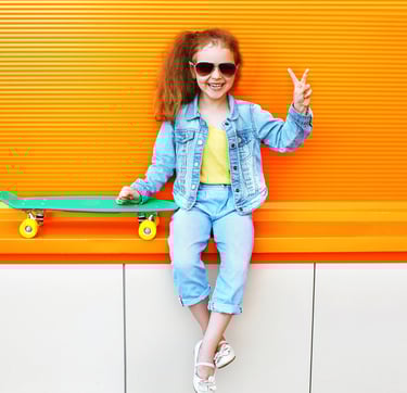 Little girl with a skateboard and holding up a peace sign, wearing kid's athletic sunglasses