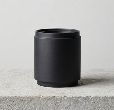 A refined still life photograph of a single charcoal black design object resting on a light gray stone surface. The background is a clean off-white wall in a Northern European studio. The lighting is precise, casting a soft medium gray shadow, embodying a modern and professional aesthetic.