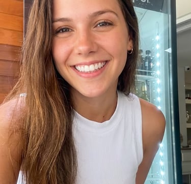 A smiling young woman with long brown hair wearing a white ribbed crop top standing in front of a cooler.