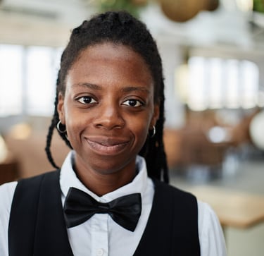 Smiling Black female server in formal uniform with a black bow tie at a modern restaurant.