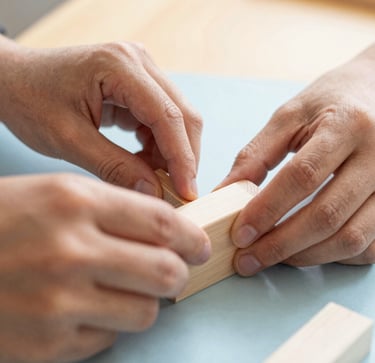 Close-up photograph of hands working on a creative school educational block project in British Columbia, bright warm background with pale blue tones.