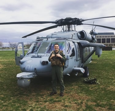 A smiling military pilot in flight gear standing in front of a gray Navy Seahawk helicopter on a grassy field.