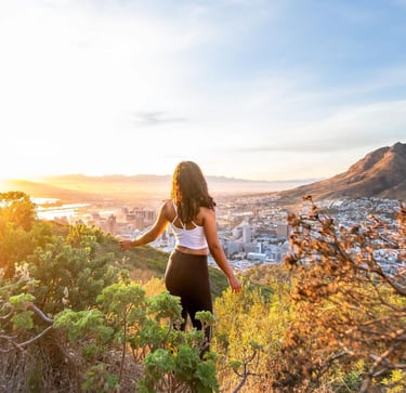 Woman standing between green bushes, looking over Cape Towns CBD. The city is basking in golden morning sunlight. 