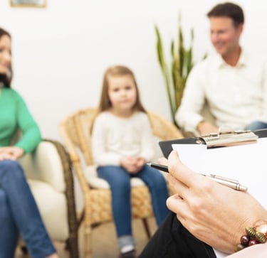 A woman sitting in a chair, holding a clipboard, surrounded by her family. Image relate to autism and ADHD in Vadodara