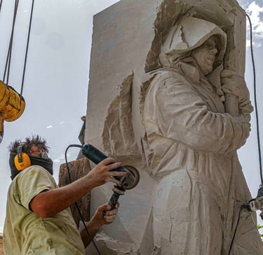 A stone sculptor using a power grinder to carve a detailed hooded figure from a large block of white marble.
