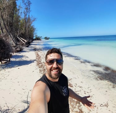 A smiling man takes a selfie on a sunny tropical white sand beach with turquoise ocean water.