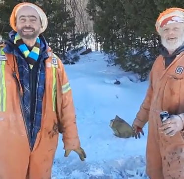 Hank & Pumpkin Head in orange safety coveralls standing in the snow near a winter forest.