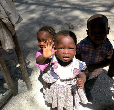 Three young African children standing outside and smiling, with one girl waving her hand.