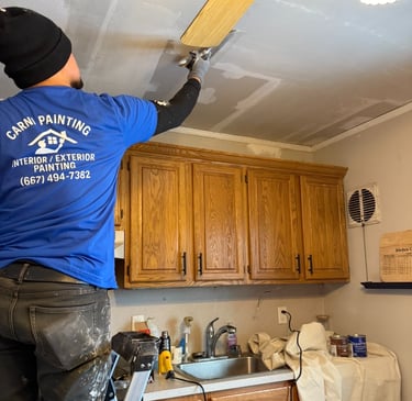 A professional painter from Carni Painting repairs and patches a kitchen ceiling above wood cabinets.