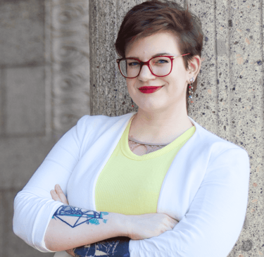 Swana Schuchmann mit Tattoos auf ihren Armen, roter Brille, gelben Top und weißem Blazer, lächelnd
