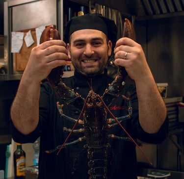 Chef Vito Amodio in cucina con divisa, sorridente mentre tiene in mano un grande astice vivo