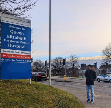 A man standing at a distance looking at the signpost reading 'Welcome to Queen Elizabeth The Queen Mother Hospital'