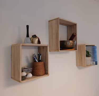 Three square wooden floating shelves on a white wall holding a singing bowl, bells, and office supplies.