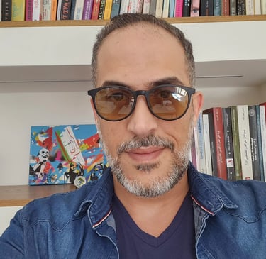 A man with a graying beard and glasses posing in front of a bookshelf with colorful books.