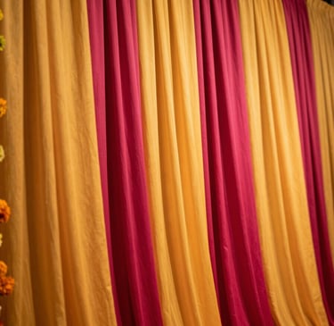Close-up of a Sangeet stage installation, vibrant marigold and fuchsia floral curtain backdrop, warm stage lighting illuminating draped fabric panels in saffron and deep magenta, wide room visible in soft focus behind
