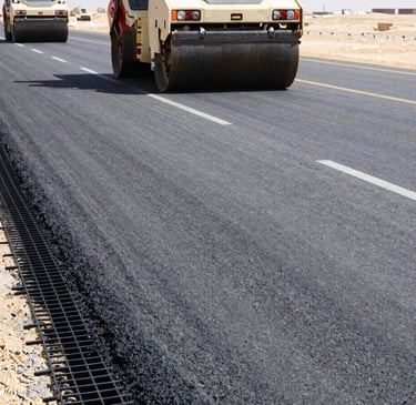 Low-angle wide shot of a multi-lane highway under construction in the Egyptian desert, black fiberglass geogrid mesh visible under a layer of fresh asphalt, heavy road rollers compacting, bright mid-day sun, industrial construction photography.
