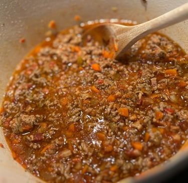 A wooden spoon stirs a savory bolognese meat sauce simmering with carrots in a white Dutch oven.