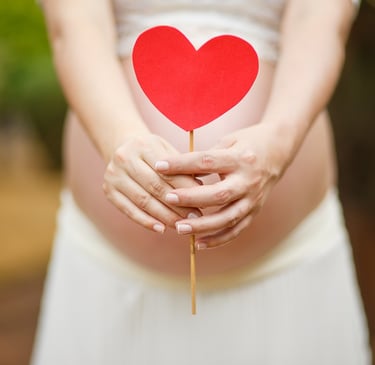 Pregnant woman holding a red heart shape over her baby bump during a maternity photoshoot.