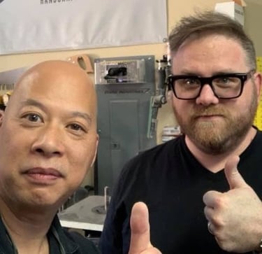 Ray Lum and Josep giving a thumbs up in a workshop setting with woodworking tools in the background.