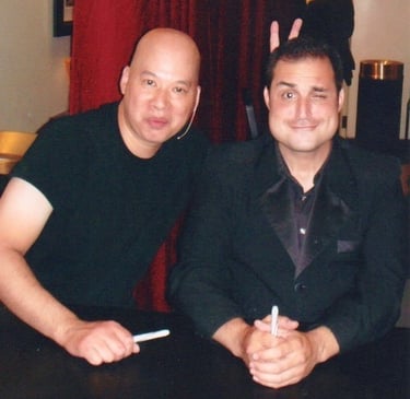 A smiling man in a black suit sitting next to a man in a t-shirt at a meet and greet event.