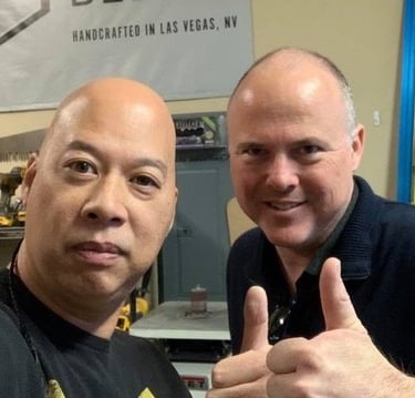 Ray and Chris giving a thumbs up in a Las Vegas woodworking shop with tools and equipment.