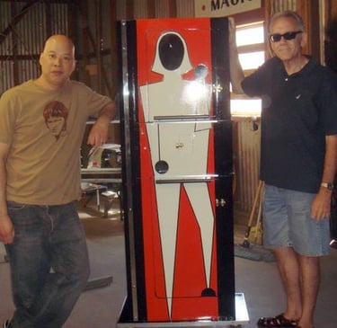 Two men standing next to a vintage Zig Zag Girl magic illusion cabinet in a workshop.