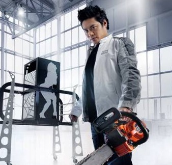 Professional magician holding an orange chainsaw next to a modern stage illusion prop in a bright studio.