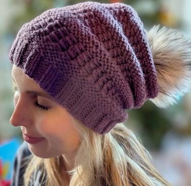 A young woman wearing a purple handknit slouchy beanie with a faux-fur pompom