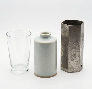A sleek, modern vase with a smooth matte finish sitting on a wooden shelf against a soft neutral wall.