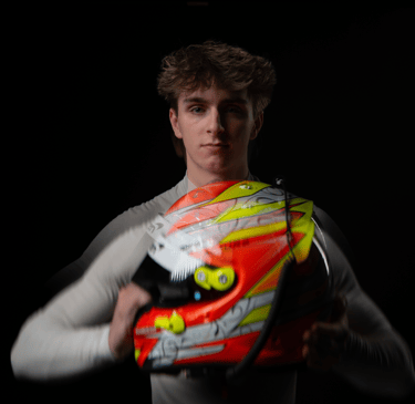 Young race car driver holding a vibrant orange and yellow racing helmet against a dark background.