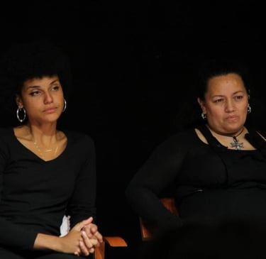 Dos mujeres actrices migrantes sentadas en el escenario en un banco de una habitación