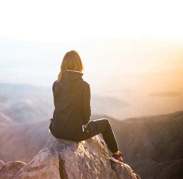 Woman sitting on mountain peak at sunrise, reflecting in solitude and enjoying peaceful nature view.