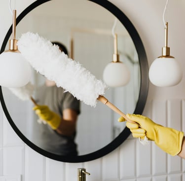 Cleaning a mirror with care — a gloved hand holding a duster in a bright, clean bathroom