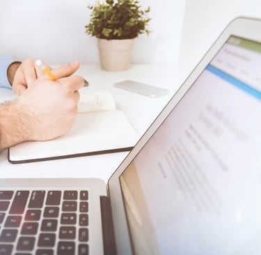 Pessoa escrevendo em caderno ao lado de notebook aberto em ambiente de trabalho