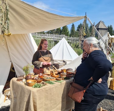a woman dressed in viking clothing standing behind a table of herbal remedies talking  with visitors