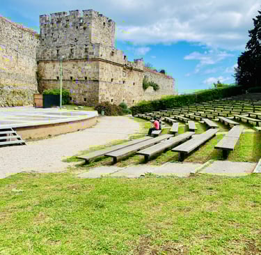 Outdoor stone amphitheater and historic castle walls in Rhodes, Greece, under a blue sky.