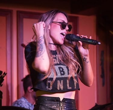 Singer Keka singing with a microphone in hand, wearing round sunglasses and a black leather skirt.