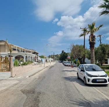 A white Kia parked on a sunny street in Greece with palm trees and Mediterranean architecture.