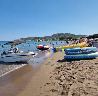 A motorboat and inflatable water sports towables sit on a crowded sandy beach under a clear blue sky.
