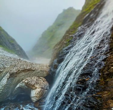 Snow-melt waterfall cascading over dark rocks in a misty mountain gorge.