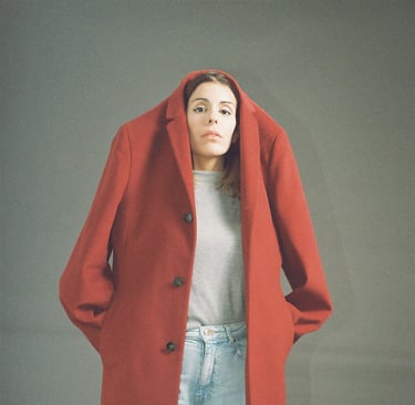 A woman wearing a large red wool coat over her head with light blue jeans against a grey background.