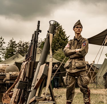 Soldat parachutiste dans un camp, bras croisés et regard au loin. Armes alignées sous un ciel sombre, tension palpable.