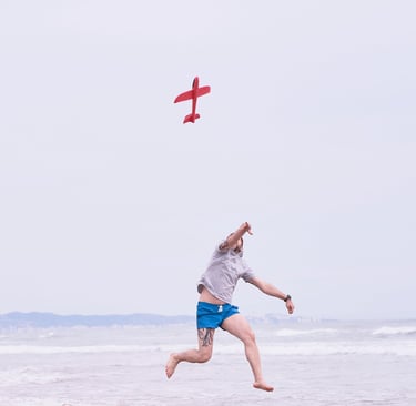 A man jumps on a sandy beach while throwing a red toy glider airplane into the air.