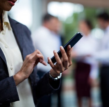 Person using a smartphone with colleagues conversing in the background.