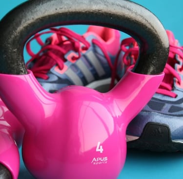 A pink kettlebell sits on a blue surface with tennis shoes in the background.