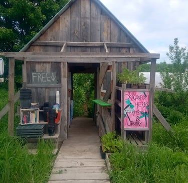 A wooden farmstand with an open entrance, featuring signs for organic eggs, free produce, and 'The D