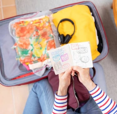 Overhead view of a person holding an open passport displaying travel stamps, next to an open suitcas