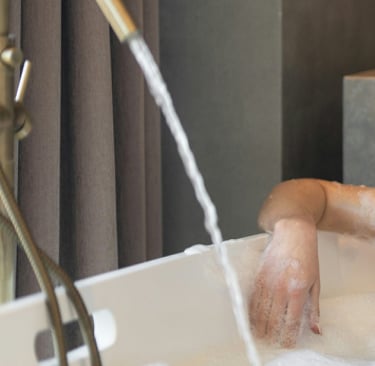 woman wearing glasses and hair wrapped in towel, relaxing in modern tub filled with water and bubbles.